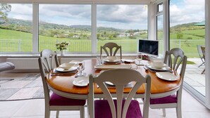 A dining area with a table set for four, large windows offering a view of green hills and a cloudy sky.