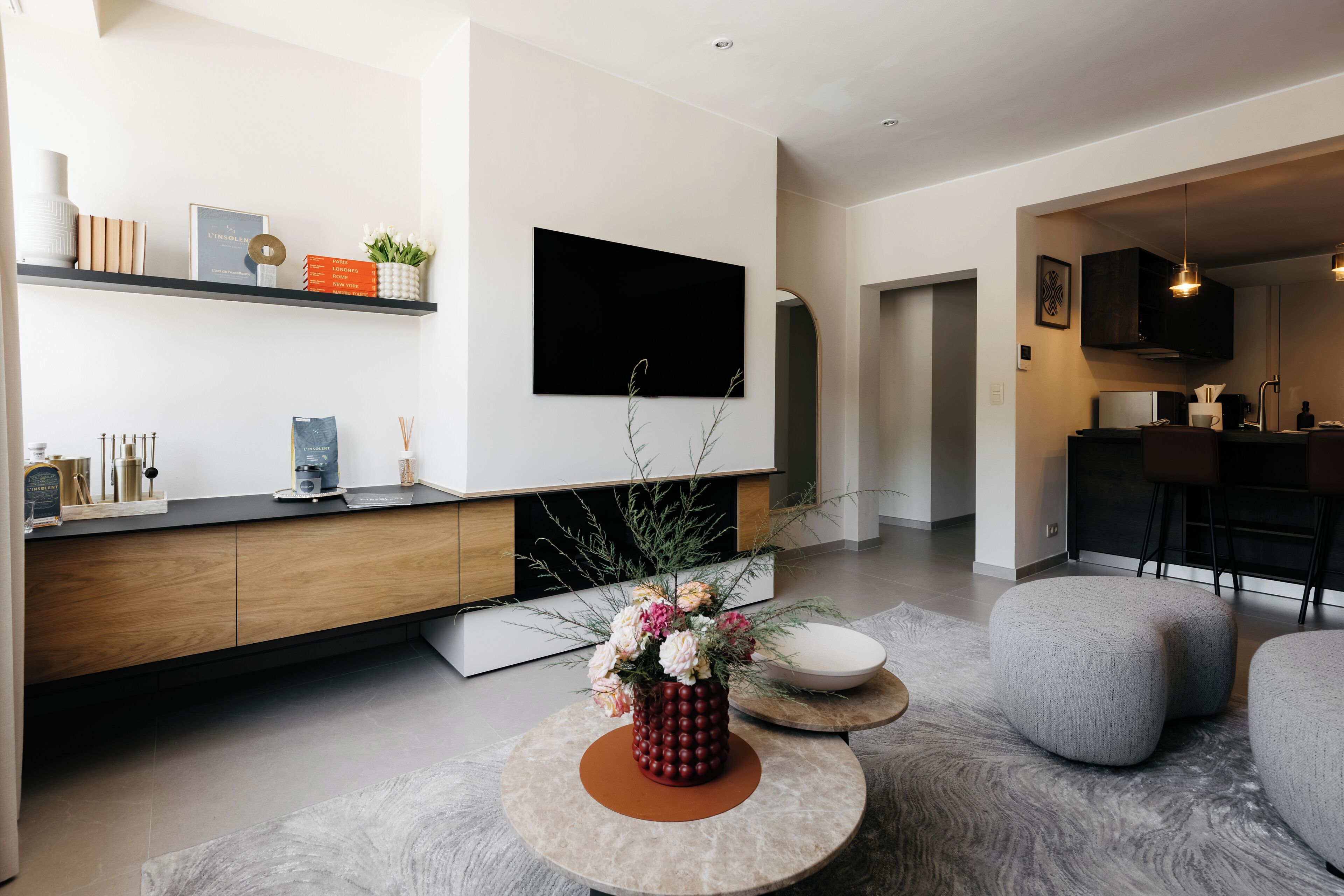 Una sala de estar moderna con un televisor de pantalla plana, una consola de madera, una mesa de centro redonda con un florero y otomanas grises.