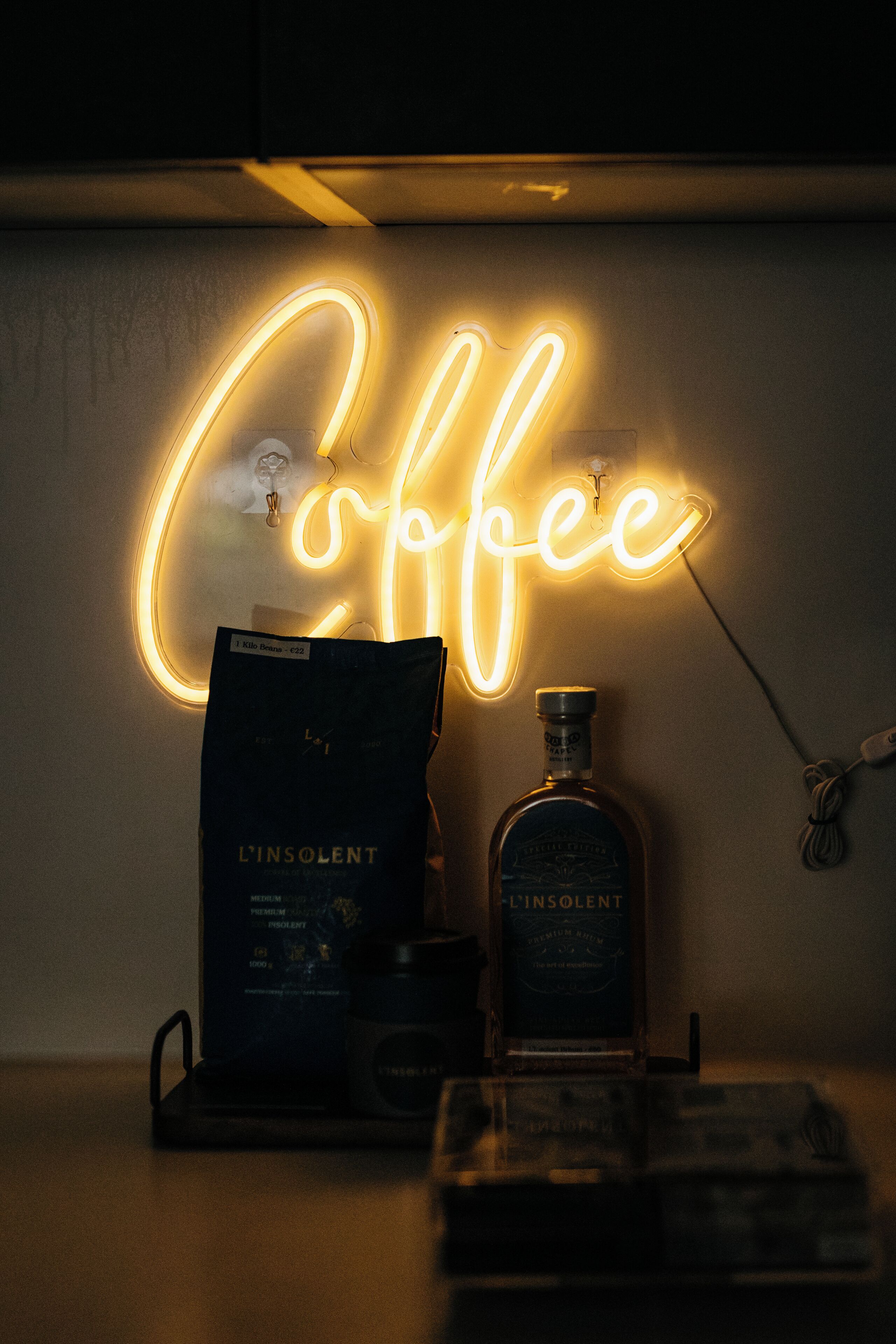En neonskilte med ordet "Coffee", en flaske L'INSOLENT spiritus, og en pose kaffebønner.