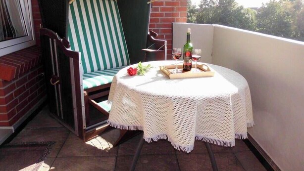 A balcony with a wicker chair, a small round table with a white crocheted tablecloth, a bottle of wine, and a glass. - Apartment Strandweh with view to the deep 2 OG-House Spiekeroog Dornumersiel (Dornumersiel)