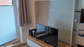 A modern room with a flat-screen TV on a wooden stand, a sliding glass door, and a window with blinds. - Apartment Strandweh with view to the deep 2 OG-House Spiekeroog Dornumersiel (Dornumersiel)