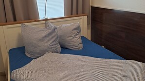 A bedroom with a bed, checkered pillows, a blue mattress, a wooden headboard, a window with curtains, and a Buddha statue on the wall. - Apartment Strandweh with view to the deep 2 OG-House Spiekeroog Dornumersiel (Dornumersiel)
