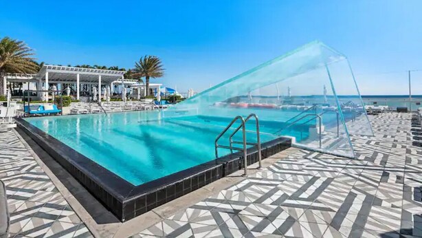 Una amplia piscina exterior con agua cristalina y azul, rodeada por una solana con baldosas y una zona de estar cubierta.