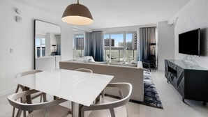 Una habitación de hotel moderna con zona de comedor, sofá, televisor y grandes ventanales con vistas a la ciudad.