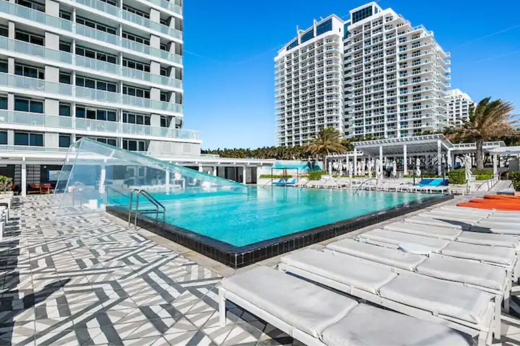 A rooftop pool with lounge chairs, a glass canopy, and high-rise buildings in the background.