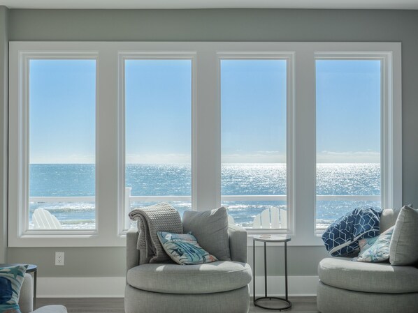 Una habitación con un ventanal grande con vista al océano, un sofá gris con cojines y una pequeña mesa redonda.