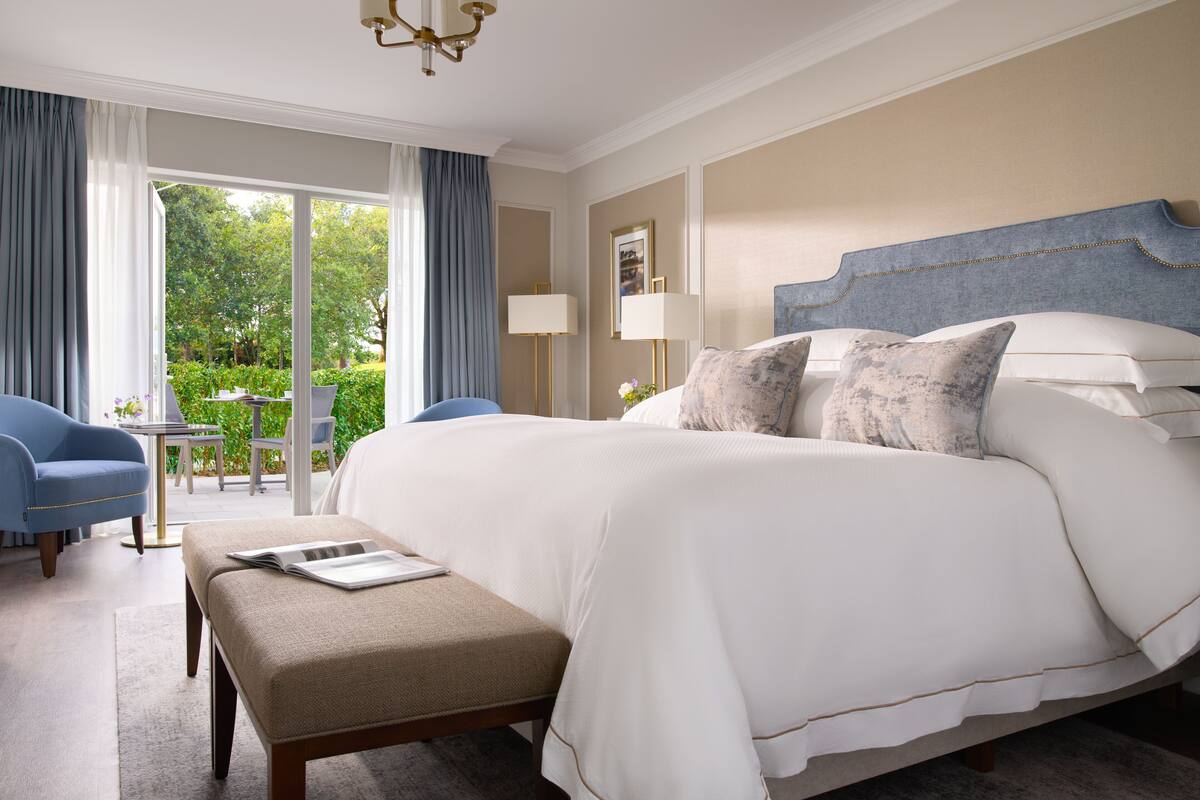 A spacious bedroom with a large bed, a small table, and a chair. There is a balcony with a view of greenery.