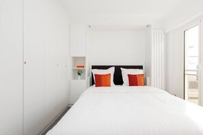 Quarto de hotel com cama, paredes brancas, cabeceira preta e uma pequena estante com uma planta.