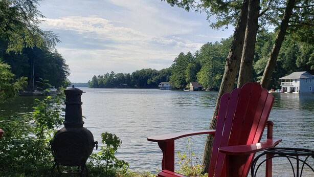 A red Adirondack chair and a grill on a stone patio overlooking a lake with trees and a house in the background.