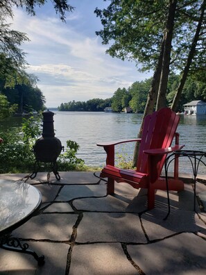 A red Adirondack chair and a grill on a stone patio overlooking a lake with trees and a house in the background.