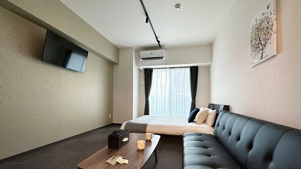 A hotel room with a bed, a sofa, a coffee table, a flat-screen TV mounted on the wall, and an air conditioning unit. - Nagomi by b hotel | Comfortable Studio Stay N41 (Hiroshima City)