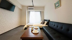 Une chambre d’hôtel comprenant un lit, un canapé noir, une table basse en bois avec des assiettes, un téléviseur à écran plat fixé au mur et un appareil de climatisation.