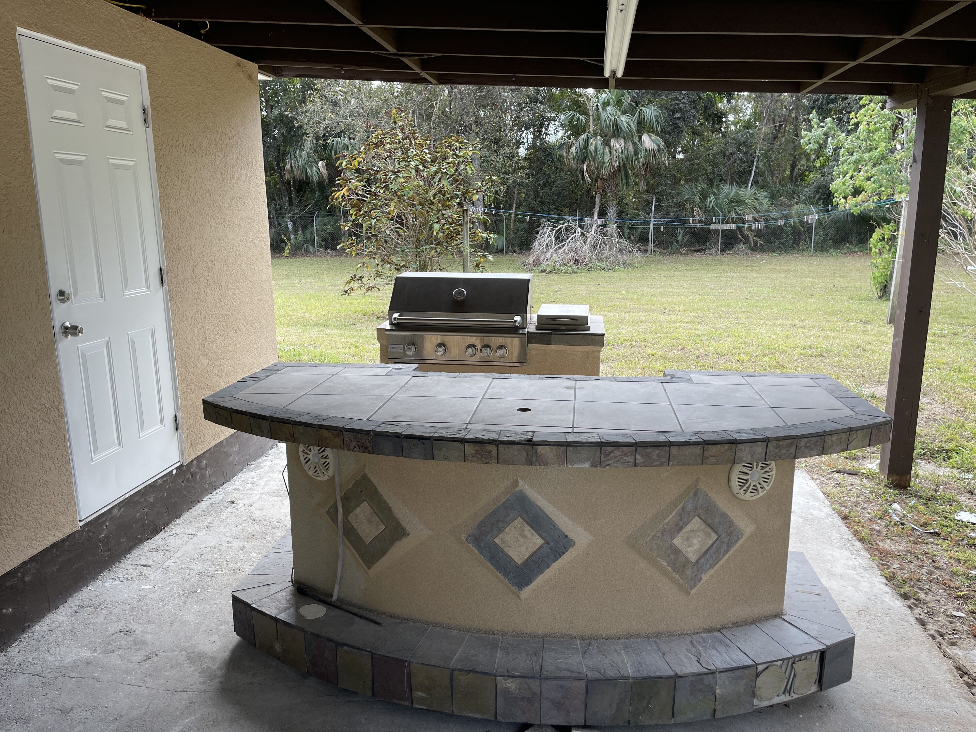 A covered outdoor barbecue area with a stone countertop and grill, surrounded by a concrete floor and a white door.