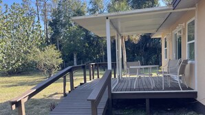 Een overdekt terras met buitenmeubilair, een houten loopplank en uitzicht op een woning.