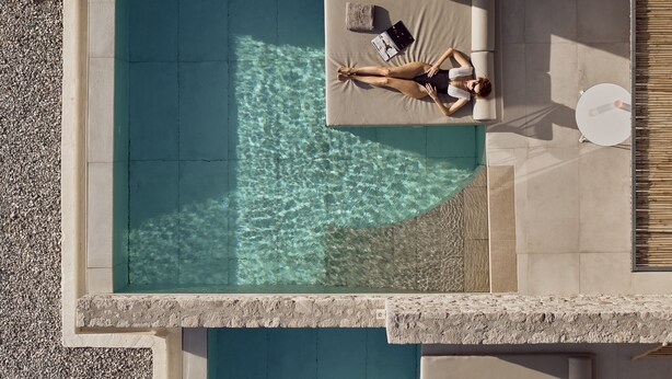 An aerial view of a swimming pool area with a lounge chair and a small table, surrounded by stone walls and a tiled deck. - Civitas Milos (Milos)