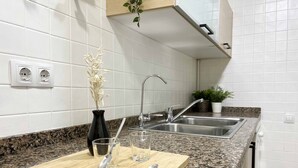 Eine Küche mit weißen Fliesen, einer Granit Arbeitsplatte, einem Holzschnittbrett, einer schwarzen Vase mit weißen Blumen, zwei klaren Gläsern, einem Löffel, einer Edelstahl-Spüle und einer Topfpflanze.