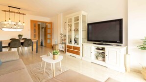Ein modernes Wohnzimmer mit Flachbildfernseher, einem Essbereich mit Stühlen, einem weißen Schrank und einem kleinen Tisch mit einer Pflanze.