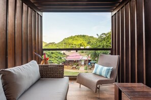 A covered patio with a sofa, a chair, and a small table. A view of a green hill and a red-roofed building is visible through the window.