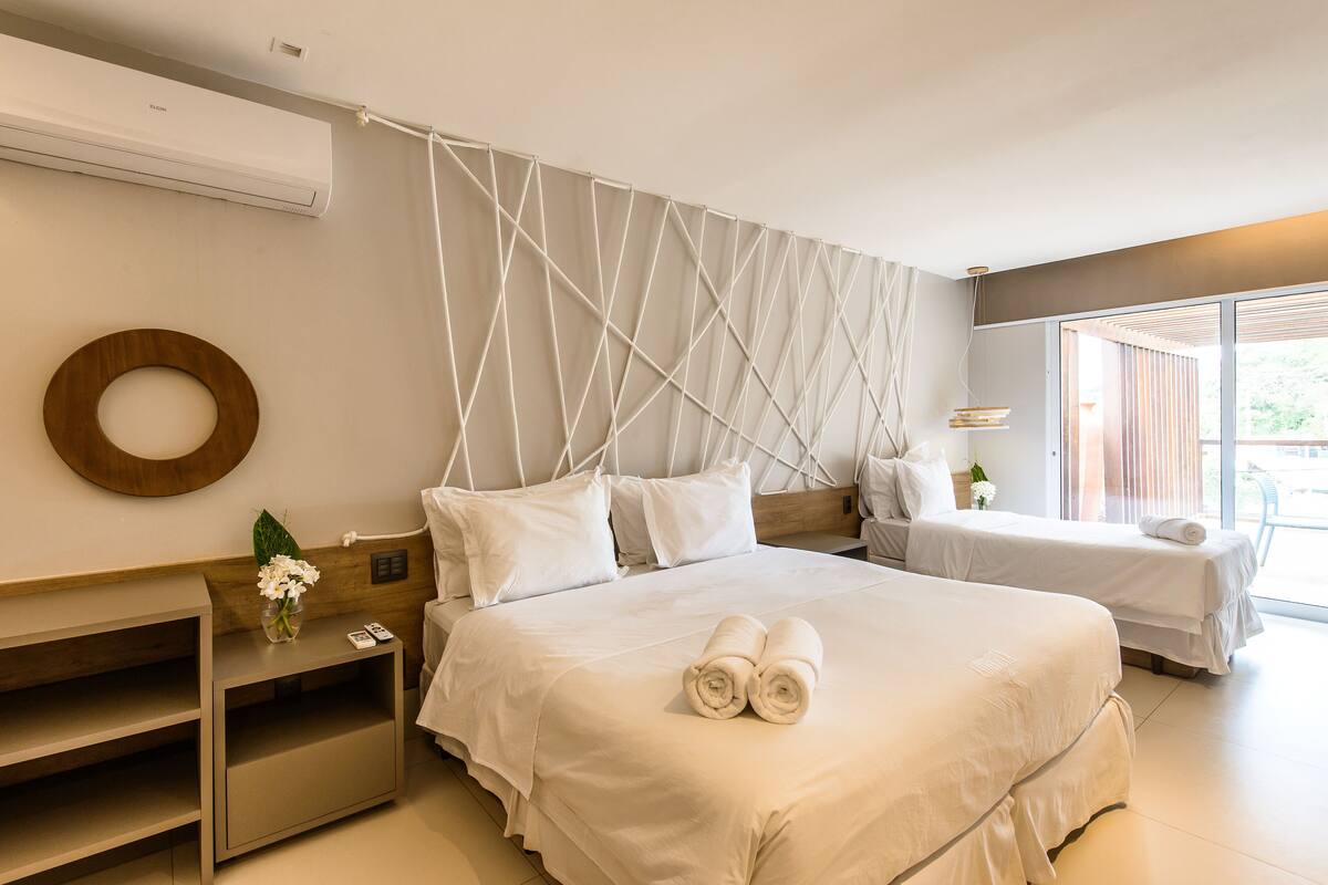 A hotel room with two beds, a wooden headboard, a wall-mounted air conditioner, a bedside table with a vase of flowers, and a view of the outdoors through large windows.