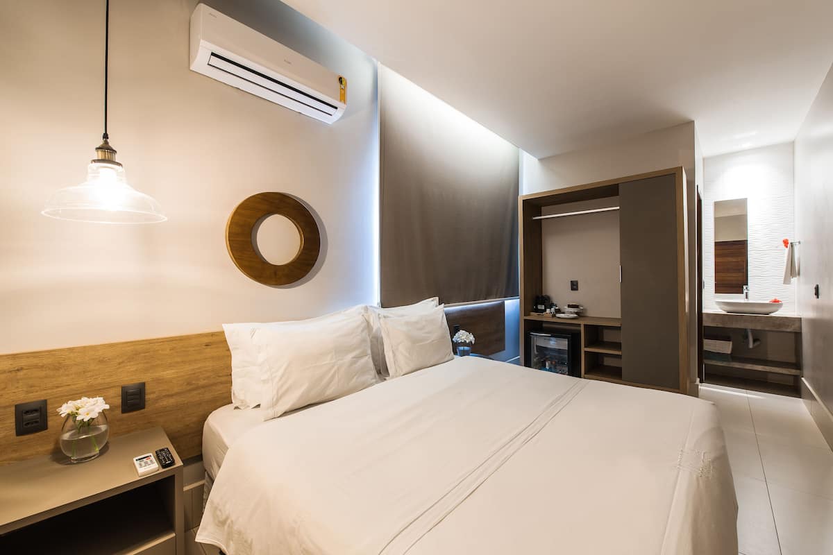 A hotel room with a bed, bedside table, lamp, air conditioning unit, and a mirror.