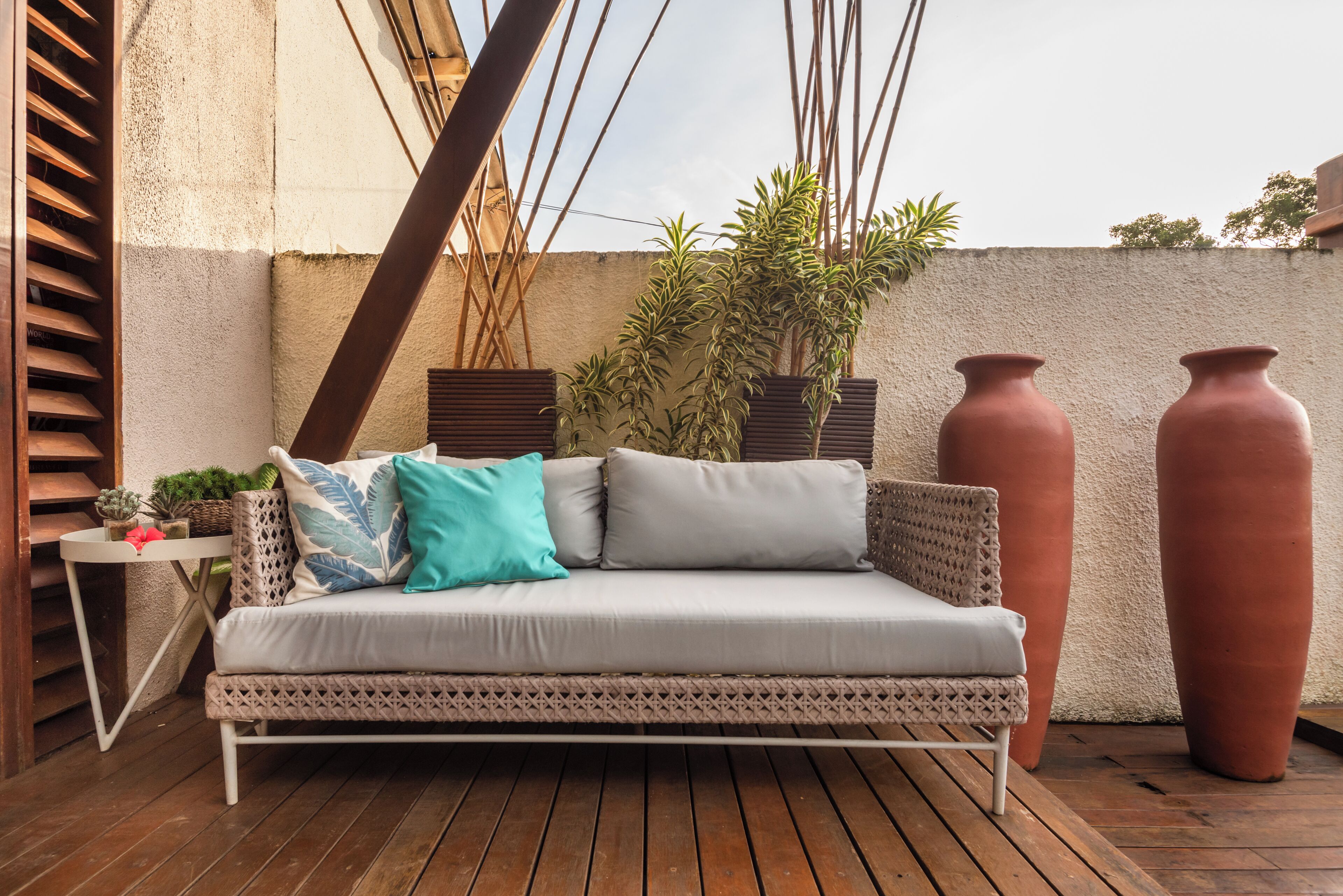 Un canapé blanc agrémenté de coussins à motifs, une petite table avec des plantes et deux grands pots en terre cuite sur une terrasse en bois.
