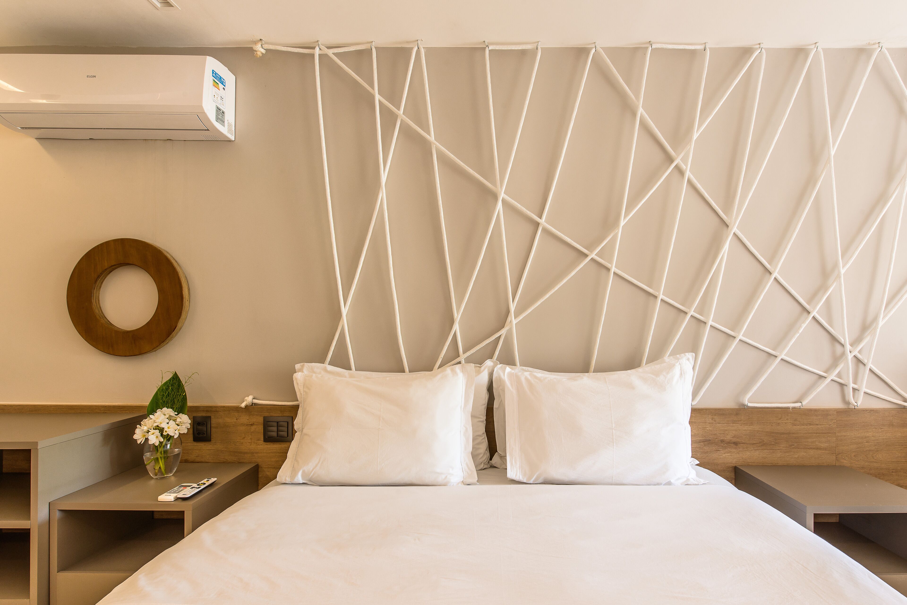 A hotel room with a bed, two pillows, a bedside table with a vase of flowers, a wooden headboard, and a wall-mounted air conditioner.