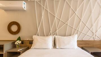 A hotel room with a bed, two pillows, a bedside table with a vase of flowers, a wooden headboard, and a wall-mounted air conditioner.