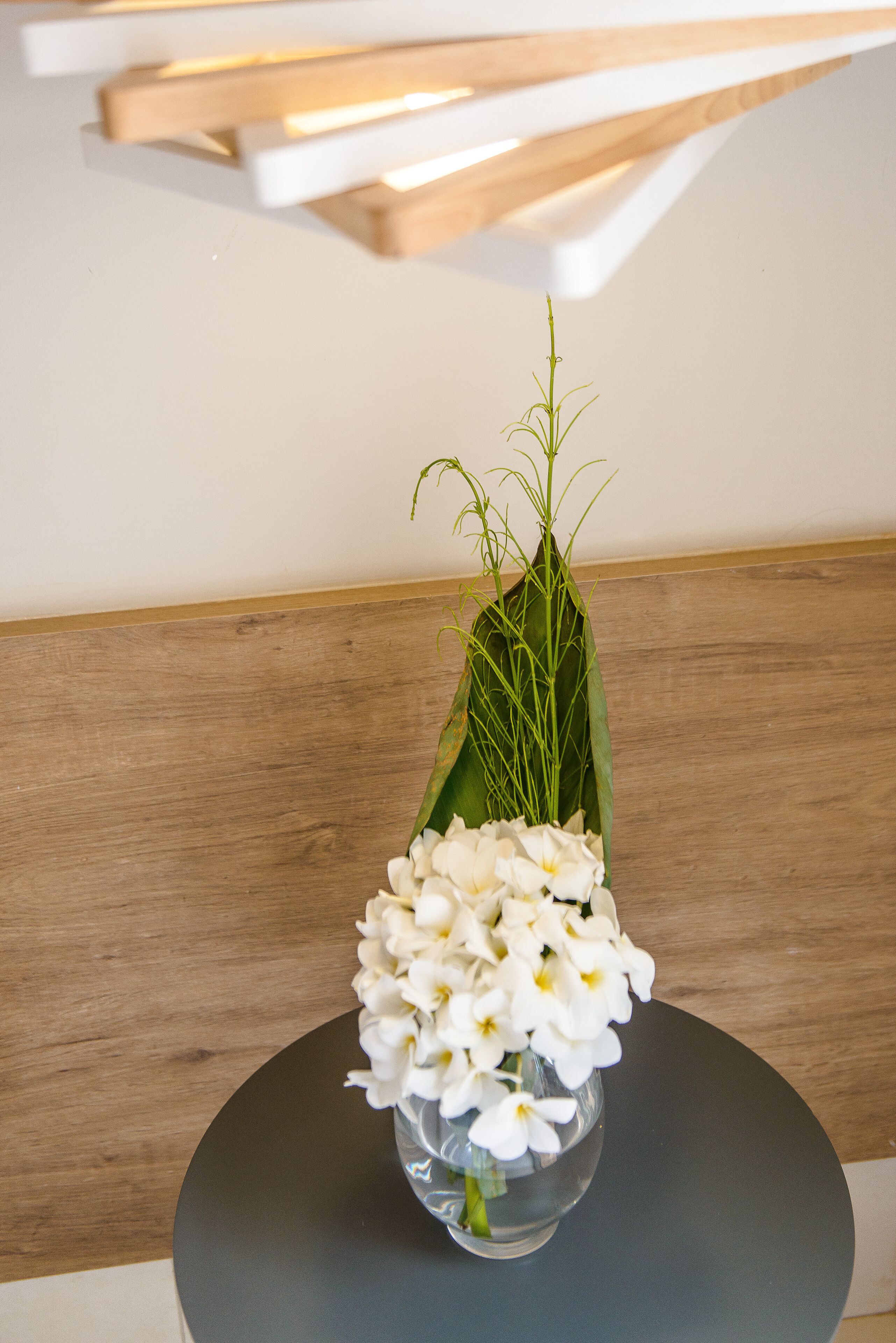 A round table with a clear glass vase containing white flowers and green foliage.