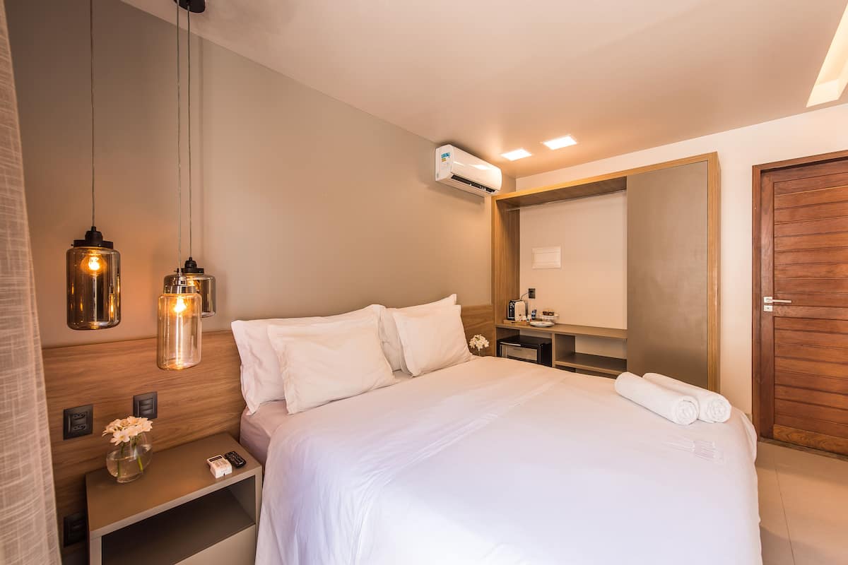 A neatly made bed with white linens, a bedside table with a vase of flowers, and a wall-mounted air conditioner.