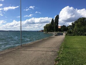 Ein gepflasterter Weg entlang eines Gewässers, mit einer Rasenfläche auf der einen Seite und Bäumen und Gebäuden in der Ferne.