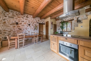 Una cocina rústica con paredes de piedra, vigas de madera, una estufa grande y un comedor con sillas y una mesa.