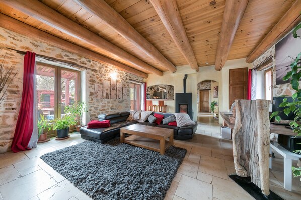 A cozy living room with a stone wall, wooden ceiling beams, a fireplace, and a large wooden table. - Villa Lorna - 2 maisons - piscine privée (Aups)