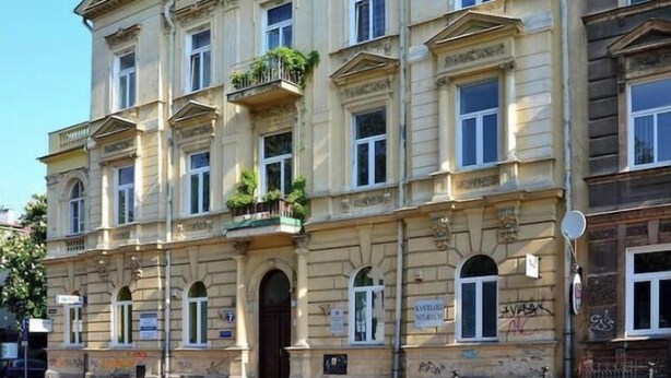 Un immeuble à étages doté d’une architecture classique, avec une entrée centrale marquée par une porte sombre, de nombreuses fenêtres et des balcons ornés de plantes.