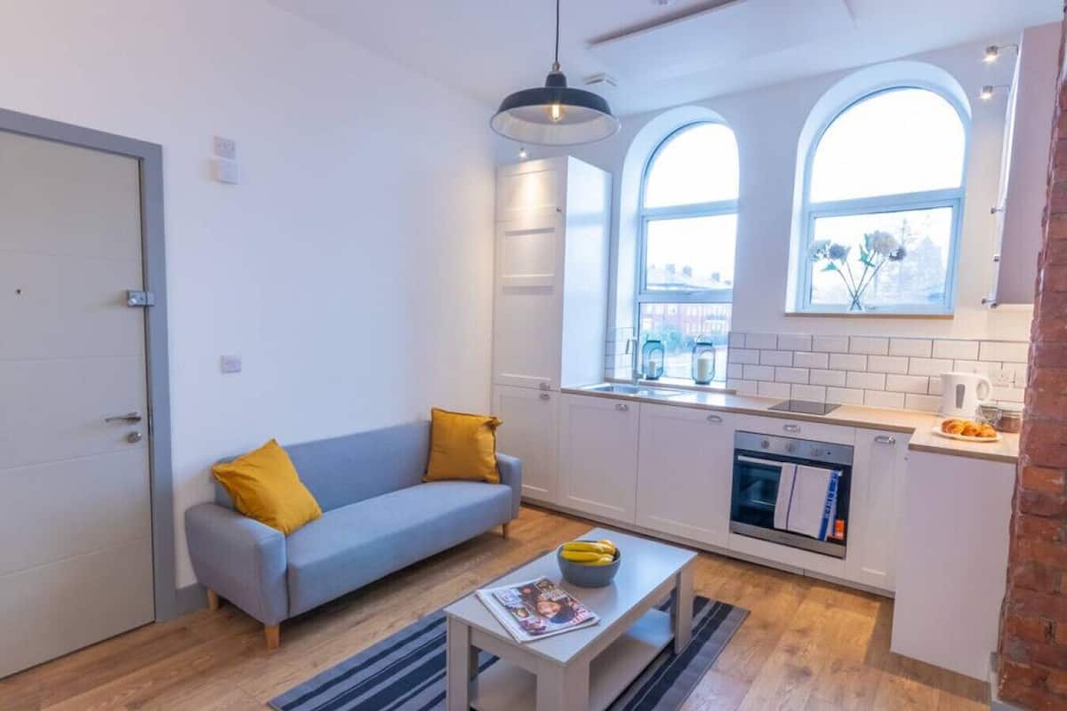 Un espacio habitable compacto con kitchenette, un sofá con cojines amarillos, una mesa de comedor con un cuenco de fruta y una ventana con vistas al exterior.