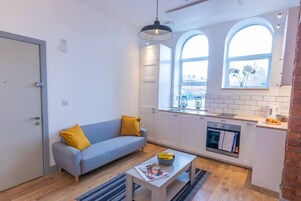 Un espacio habitable compacto con kitchenette, un sofá con cojines amarillos, una mesa de comedor con un cuenco de fruta y una ventana con vistas al exterior.