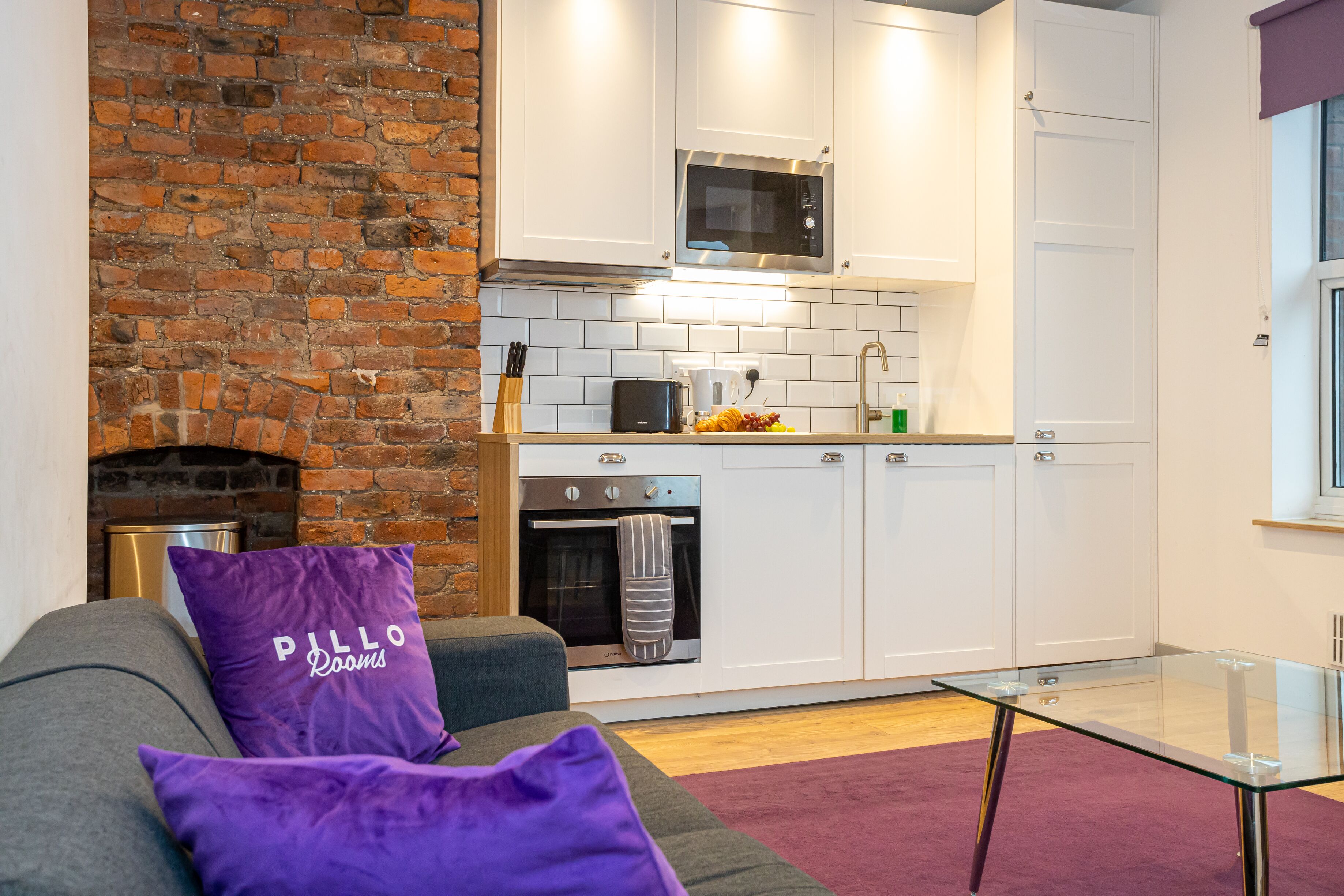 A modern kitchen with white cabinets, a brick wall, a microwave, a toaster, and a glass table.