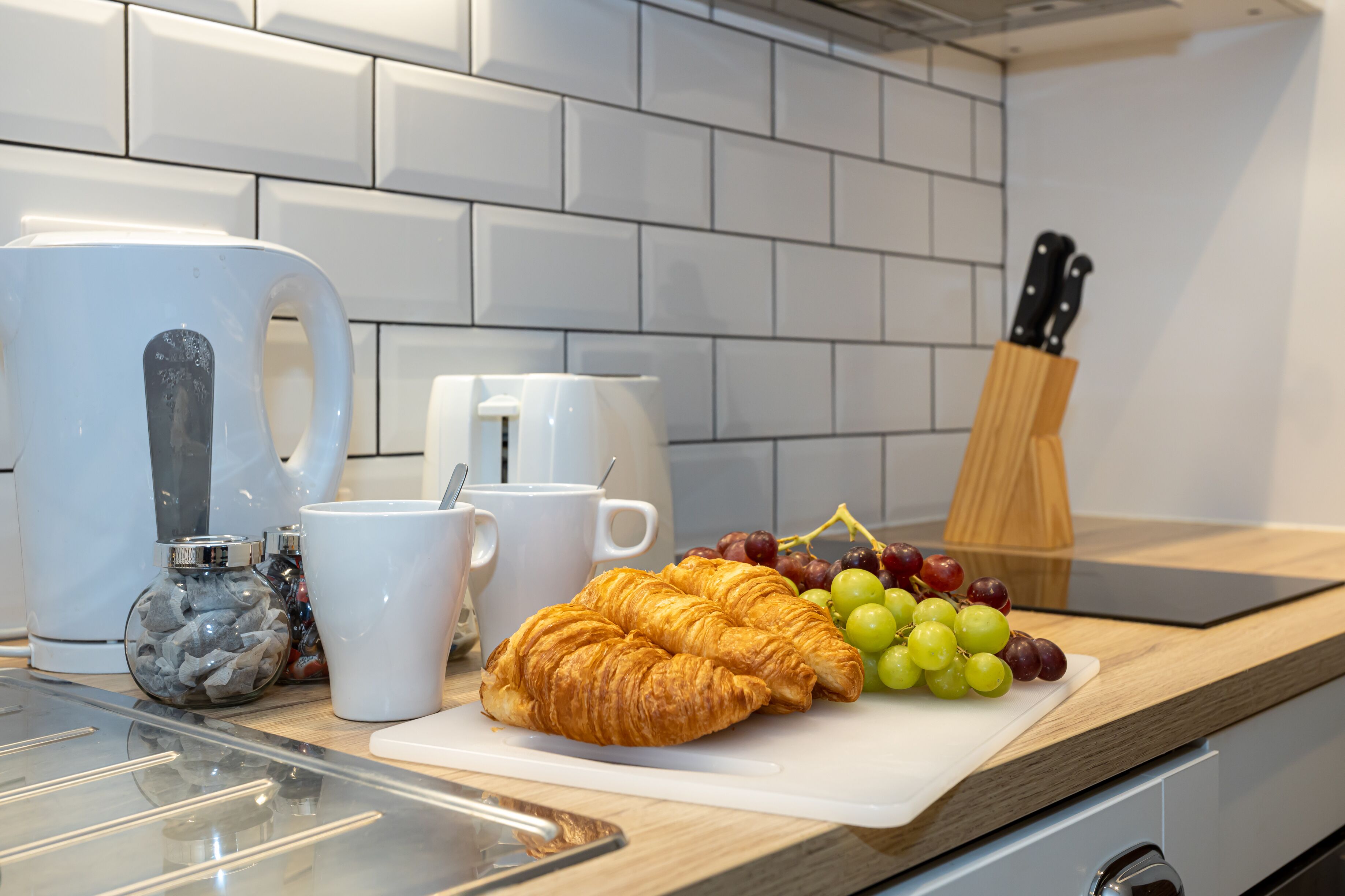 A kitchen counter with a cutting board, two croissants, a bunch of grapes, a coffee maker, and a knife block.