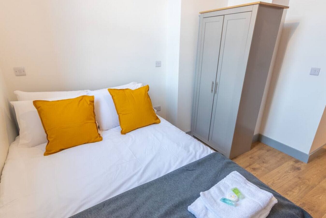 A bed with white bedding and two mustard pillows, a grey blanket, and a small white towel with toiletries on a wooden floor.