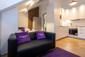 Un salon moderne avec un canapé en cuir noir, deux coussins violets et un tapis violet. À côté se trouve une cuisine avec des armoires blanches, un mur en briques et un four encastré.