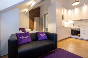 Una sala de estar moderna con un sofá de cuero negro, dos almohadas moradas y una alfombra púrpura. Junto a ella, una cocina con gabinetes blancos, una pared de ladrillo y un horno empotrado.