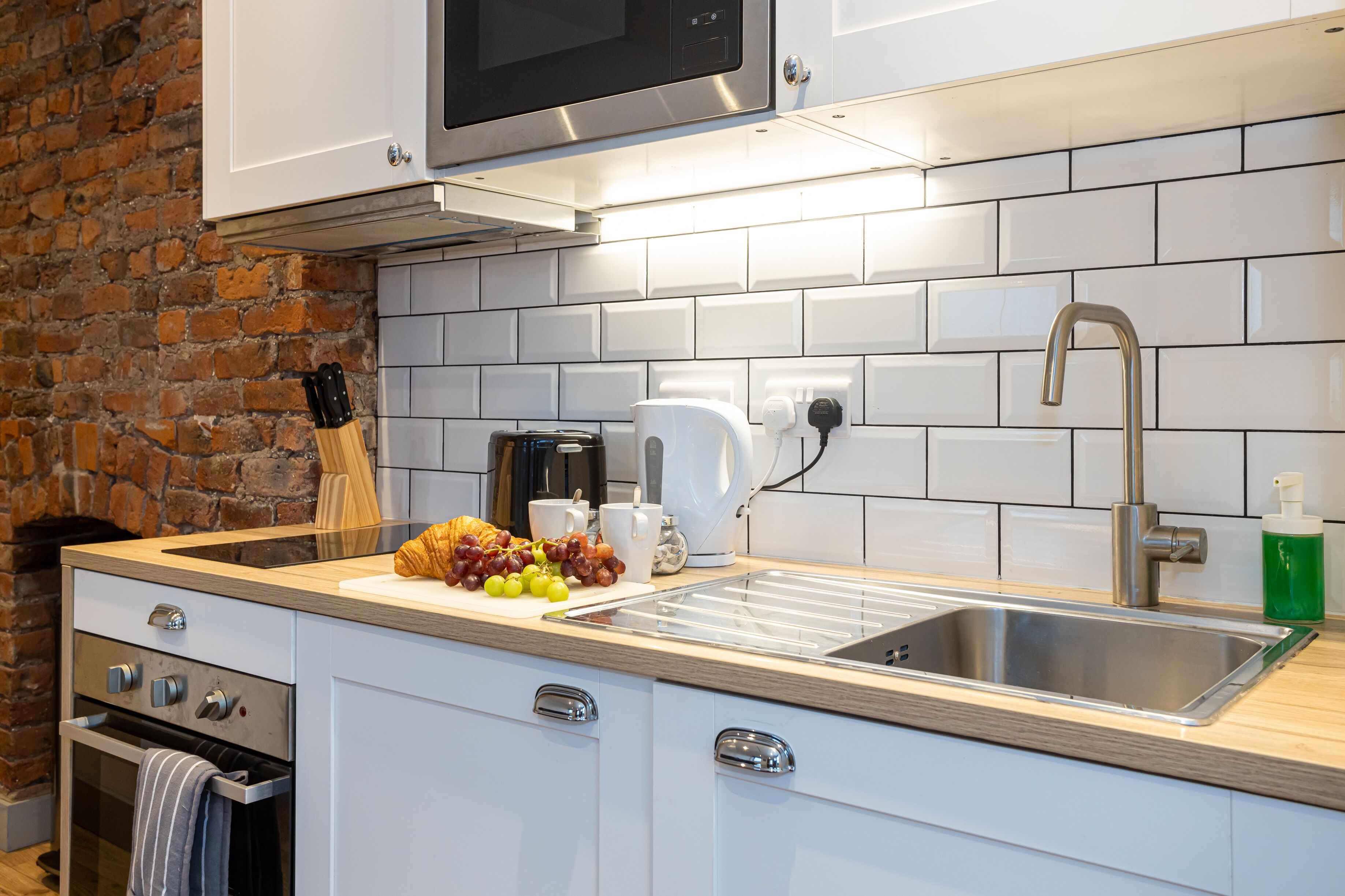 Una cucina moderna con mobili bianchi, un lavello in acciaio inossidabile, un forno a microonde e una parete in mattoni.