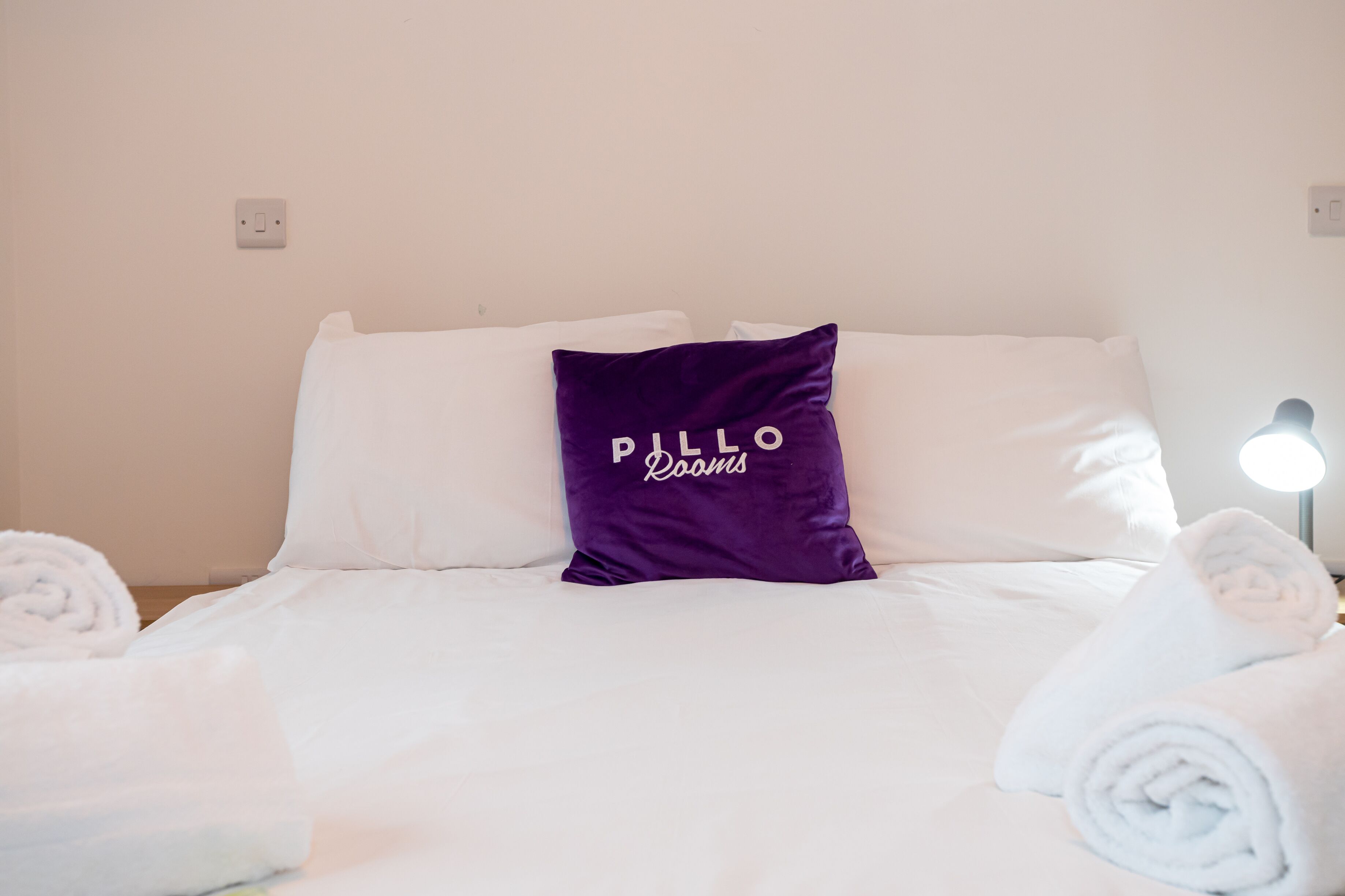 Een net opgemaakt bed met witte lakens, een paars kussen met de tekst 'PILLO Rooms' en witte handdoeken opgerold aan weerszijden.