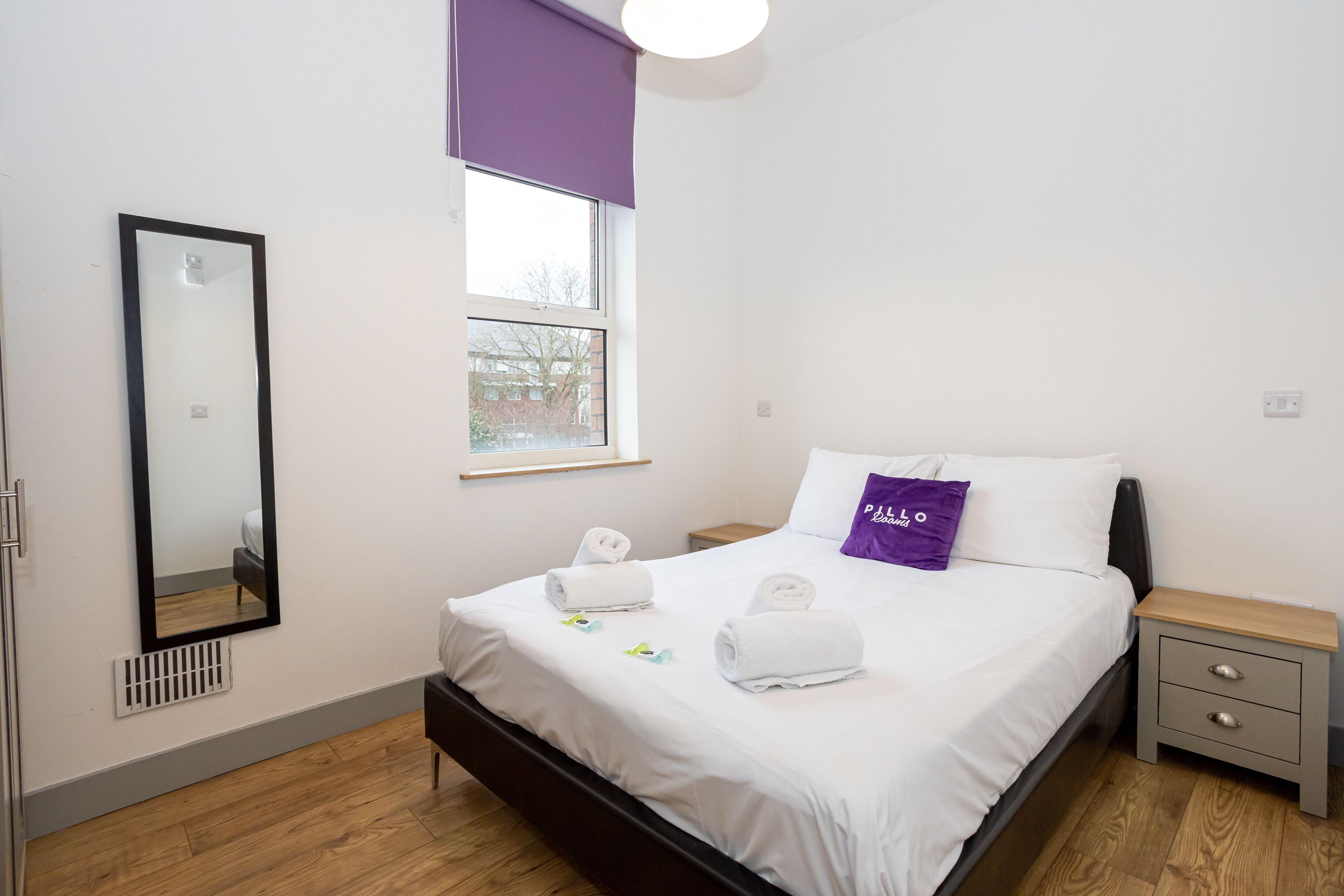 Una cama bien hecha con sábanas blancas, una almohada morada y dos toallas enrolladas. Un espejo grande cuelga de la pared, y se ve una ventana con vistas a edificios.