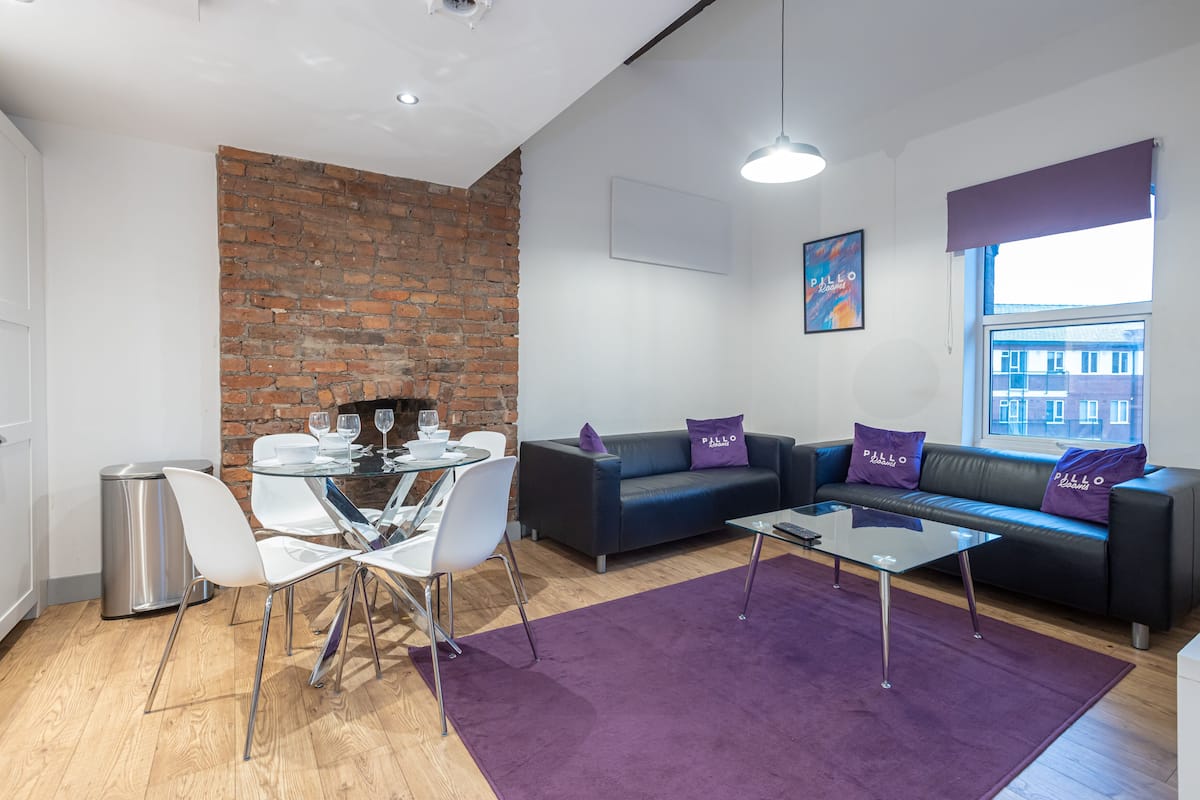 Una sala de estar moderna con una pared de ladrillo, un sofá con cojines morados, una mesa de comedor de cristal y una alfombra morada.