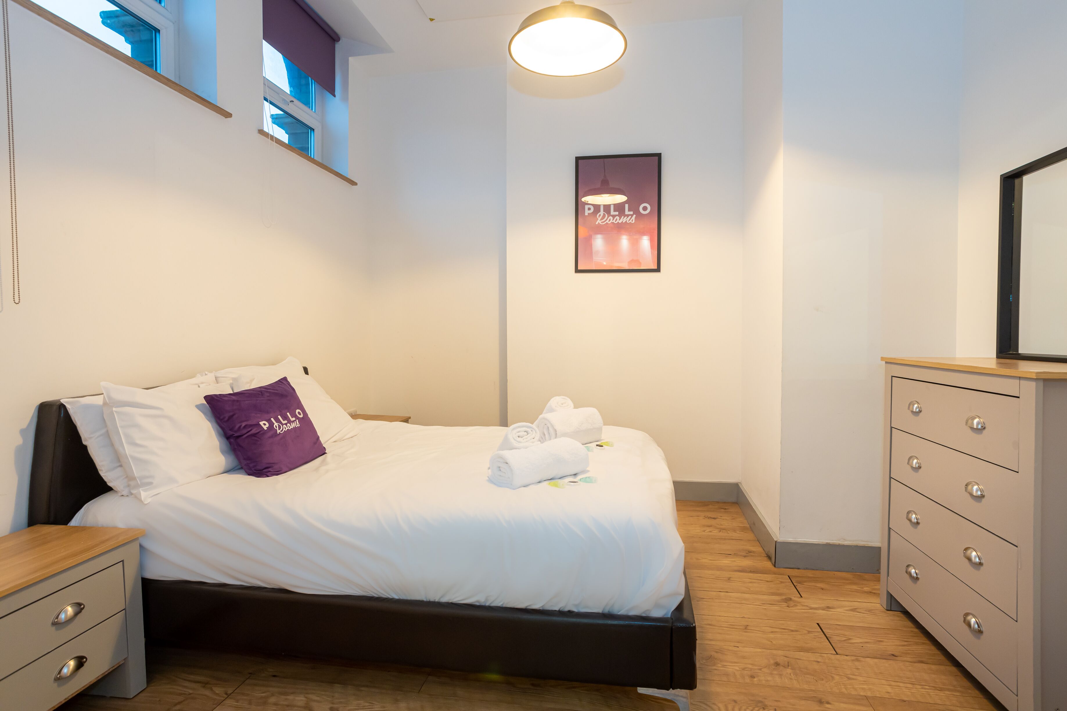 A neatly made bed with white bedding and a purple pillow, a wooden nightstand, a dresser with drawers, and a framed picture on the wall.