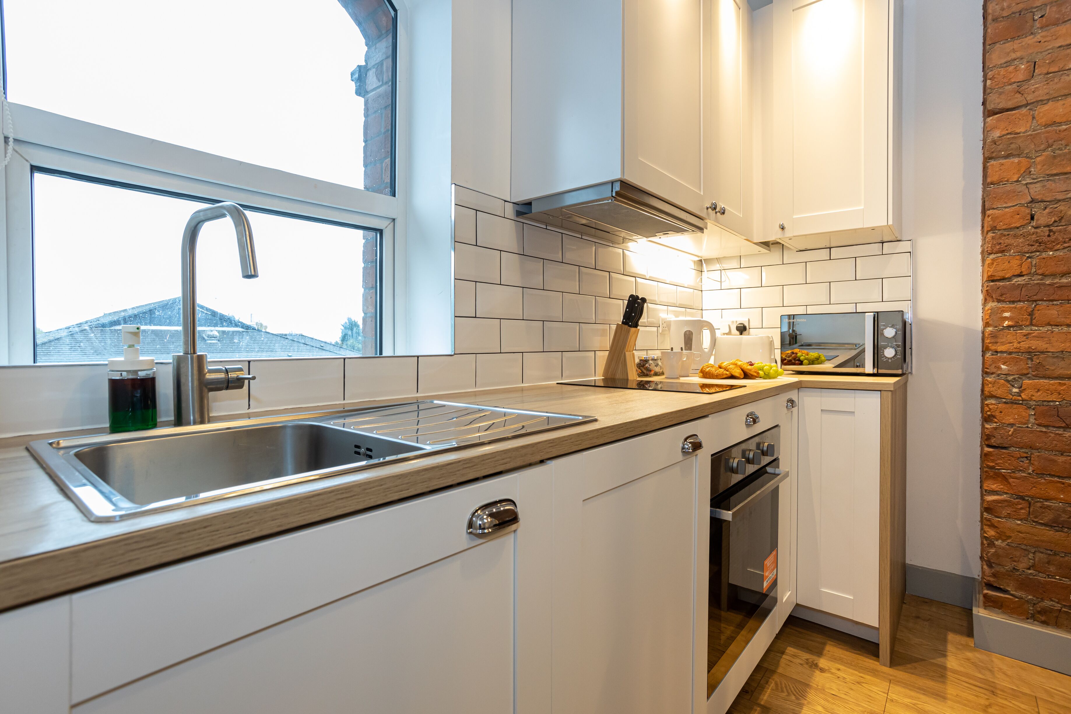 A modern kitchen with white cabinets, a stainless steel sink, a built-in oven, and a brick accent wall.