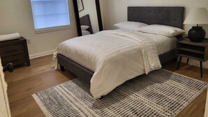 A bedroom with a bed, a nightstand, a lamp, a mirror, and artwork on the wall. - Upscale & Elevated - 3BR 2BA (Newark)