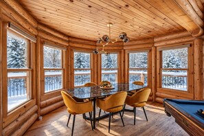 Interno di una baita di legno con tavolo da biliardo, tavolo da pranzo con sedie e ampie finestre che si affacciano su un paesaggio innevato.