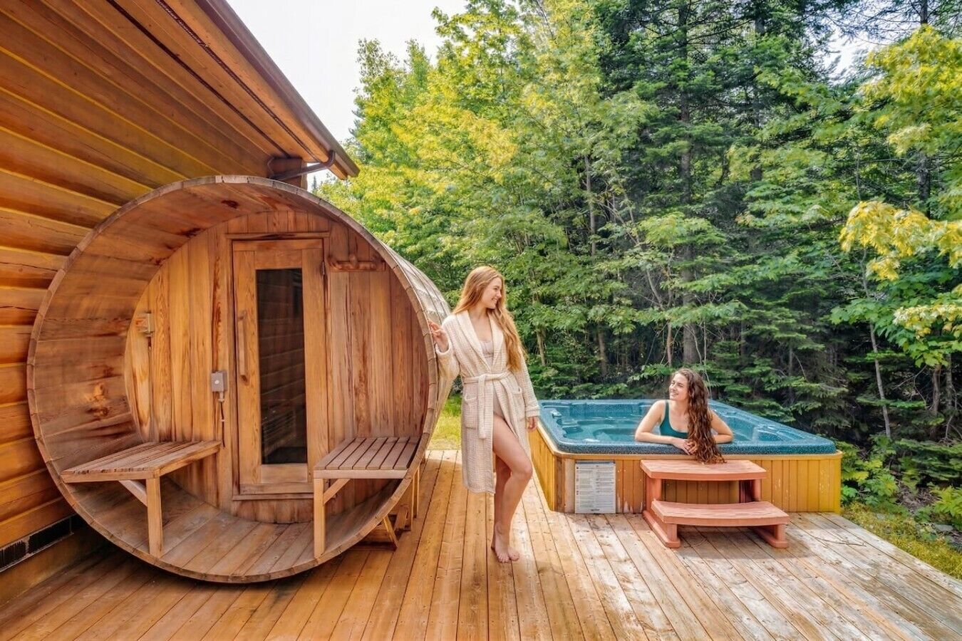 A wooden cabin with a sauna and a hot tub on a wooden deck surrounded by trees.