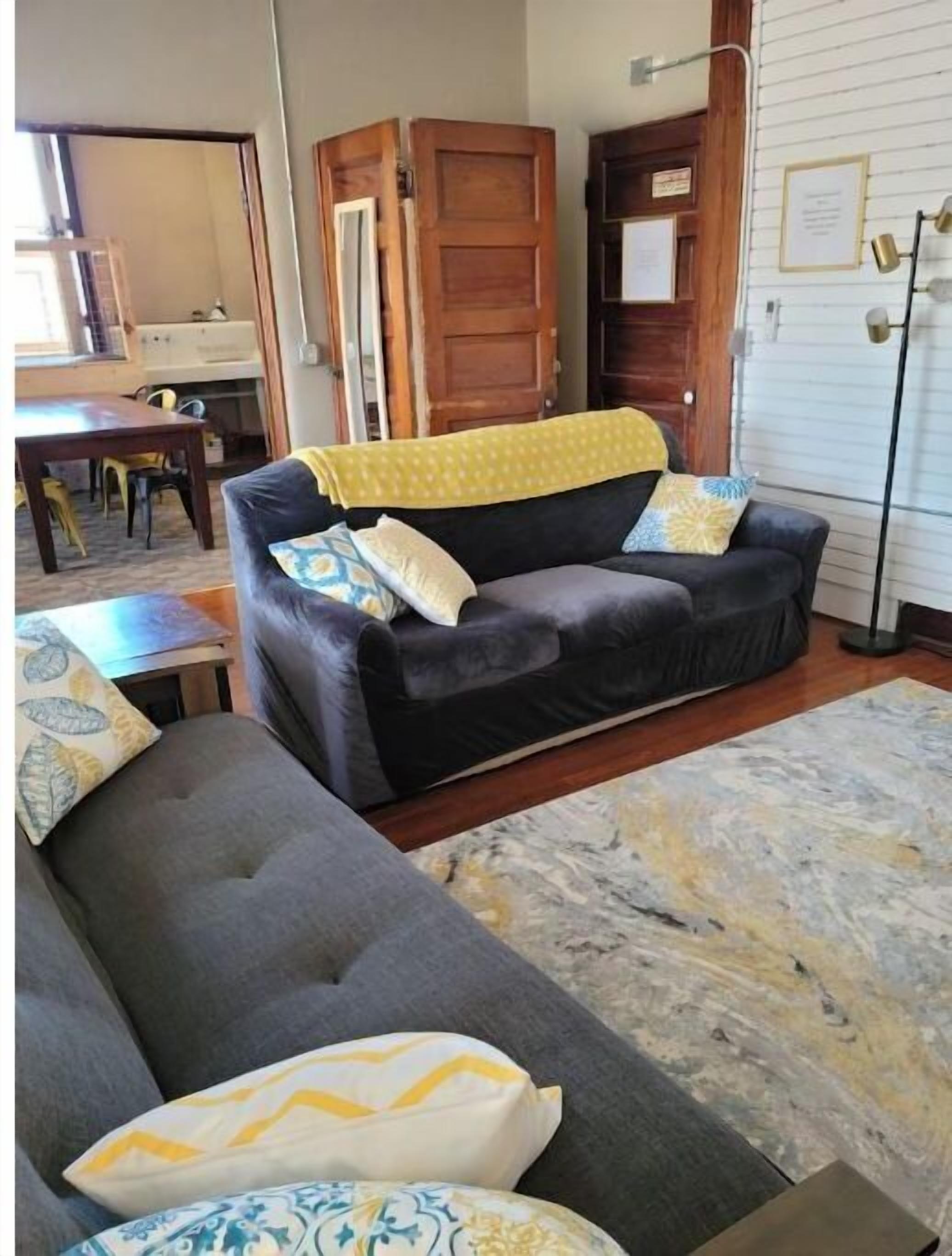 A living room with a dark grey sofa, a yellow throw blanket, and patterned cushions. There is a wooden cabinet and a floor lamp.